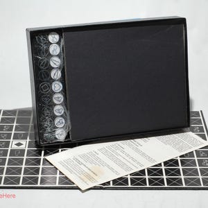 May include: A black box game with a black and white game board, a set of white and black game pieces, and a set of instructions. The game board is made of a thick cardboard and has a grid pattern. The game pieces are made of plastic and have different shapes and sizes. The instructions are printed on a sheet of paper.