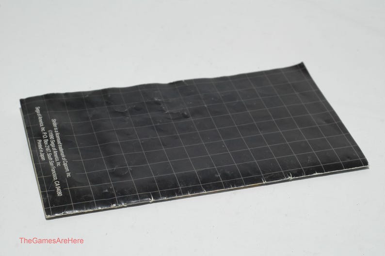 May include: A black, rectangular game manual with a grid pattern and white text. The manual is slightly crumpled and lies on a white surface. Text includes the Sega of America address.