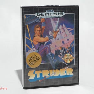 May include: A black Sega Genesis game cartridge case featuring the game "Strider." The cover art depicts a muscular man wielding a sword, with the Sega Genesis logo at the top. The game title is in a blue and white font.