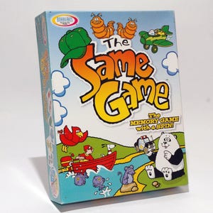 May include: A colorful box for "The Same Game," a memory game with a spin. The box features cartoon illustrations of a red bird, a panda, and other whimsical characters. The game's title is in large, orange and yellow letters.