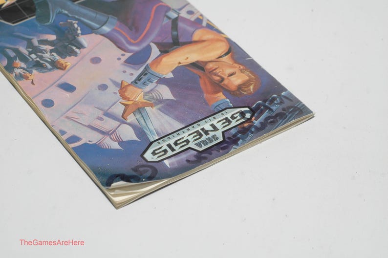 May include: A Sega Genesis 16-bit cartridge game box. The box art features a muscular man holding a sword, with a futuristic background. The Sega Genesis logo is prominently displayed. The box is on a white surface.