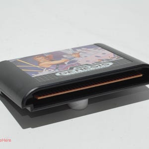 May include: A black Sega Genesis video game cartridge. The cartridge has a colorful label with a graphic of a person and the Sega Genesis logo. The cartridge is resting on a small, silver stand.