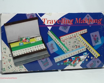May include: A blue box with the words "Traveling Mahjong" on the front. The box contains a set of Mahjong tiles, dice, and other accessories. The tiles are arranged in rows and columns, and the dice are in a small plastic container. The box is open, and the tiles are visible.