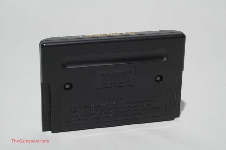 May include: Black SEGA Genesis game cartridge. The cartridge is rectangular with rounded edges. The SEGA logo is visible. The text "TheGamesAreHere" is at the bottom of the image.