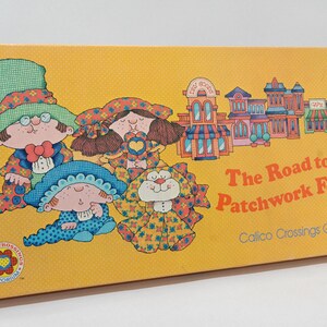 May include: A yellow box with a game called "The Road to the Patchwork Forest" Calico Crossings Game. The box features illustrations of four children and a cat wearing colorful patchwork clothing. The children are wearing hats and the cat has a bow tie.