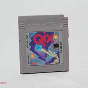May include: A gray Nintendo Game Boy cartridge for the game Qix. The label features the game's title in red and a geometric design with blue and yellow accents. The cartridge is marked "DMG-OX-USA" and "Made in Japan."