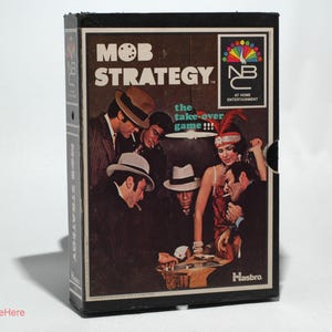 May include: A box for the board game "Mob Strategy: The Take-Over Game!" by Hasbro. The box features a group of people in 1920s attire, including a woman in a flapper dress and a man in a fedora. The box is black with a white and red design.