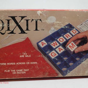 May include: A QIXIT board game box featuring a blue grid tray with lettered cubes spelling A WORD GAM. The box includes a red triangular area with text stating X'S ARE WILD, FORM WORDS ACROSS OR DOWN, and PLAY THE GAME FAST... OR FASTER!