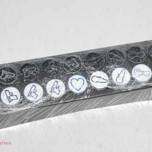 May include: A black plastic case with a clear plastic cover containing 14 round black and white stickers with various symbols and designs. The symbols include a gun, a heart, a car, and a key.