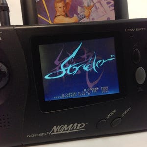 May include: A black Sega Genesis Nomad handheld game console with a Strider game cartridge inserted. The game screen displays the Strider logo in blue and purple. The cartridge features a muscular man with a sword.