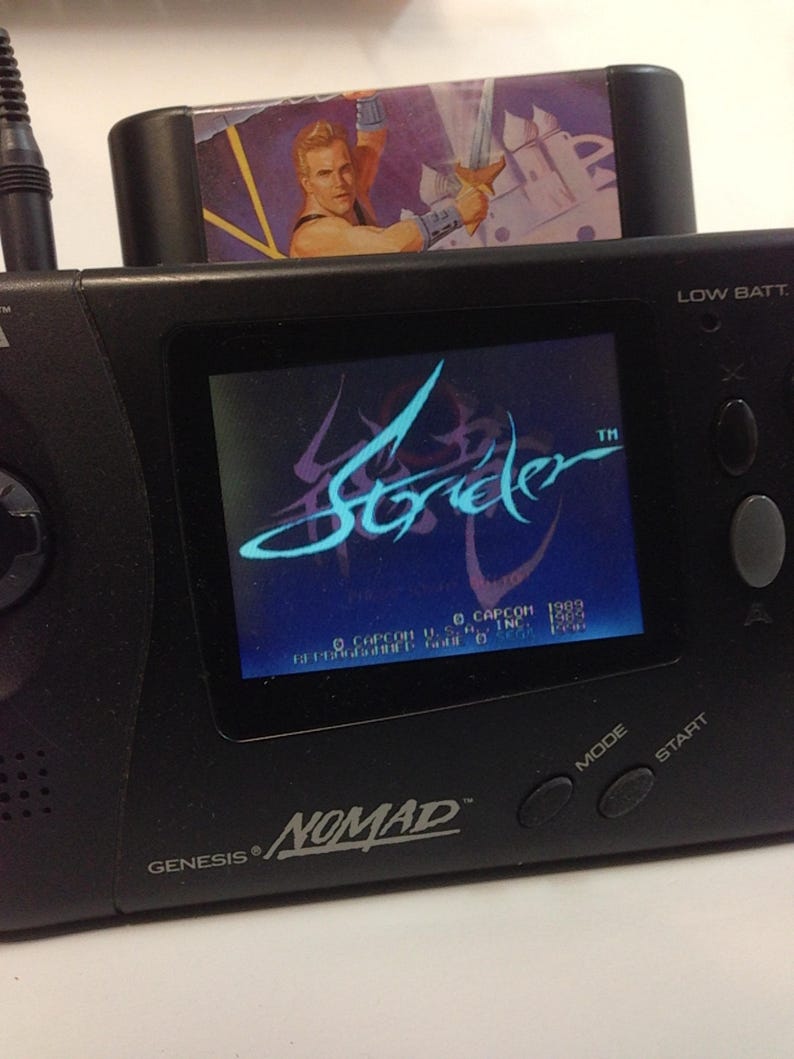 May include: A black Sega Genesis Nomad handheld game console with a Strider game cartridge inserted. The game screen displays the Strider logo in blue and purple. The cartridge features a muscular man with a sword.