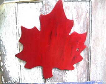 May include: A red wooden maple leaf cutout, painted in a distressed style, is mounted on a white wooden surface.