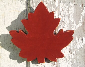 May include: A red wooden maple leaf cutout, painted in a deep red color, with a slightly rough texture. The leaf is attached to a white, weathered wooden surface.
