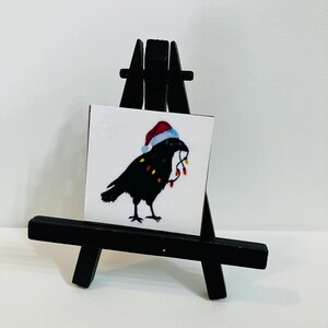 May include: A black crow wearing a Santa hat and holding Christmas lights in its beak, displayed on a small black easel. The crow is silhouetted against a white background, creating a festive holiday scene.