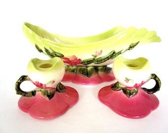 May include: A vintage ceramic centerpiece set with a green and white bowl and two matching candle holders. The bowl and candle holders are decorated with pink and green floral designs.