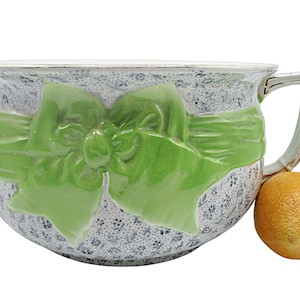 May include: A white ceramic chamber pot with a green ribbon bow design. The pot has a handle and a floral pattern.