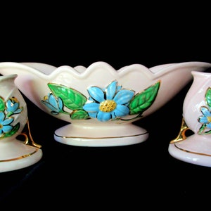 May include: A set of three vintage ceramic vases with a pink base and gold accents. The vases feature a floral design with blue and green flowers.
