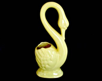 May include: A yellow ceramic swan-shaped vase with a textured surface. The vase has a wide opening at the top for flowers or other decorative items.