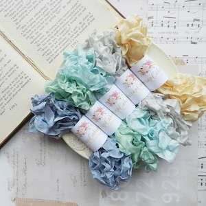 May include: Five spools of colorful silk ribbon, each wrapped in white paper with a floral design and the text "Wrap Cabin Scrap Cabin". The ribbons are in shades of blue, green, yellow, and gray.