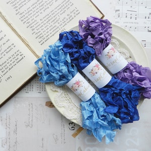 May include: Three spools of ribbon in shades of blue and purple. The ribbon is ruffled and has a satin finish. The spools are labeled "Scrap Cabin".