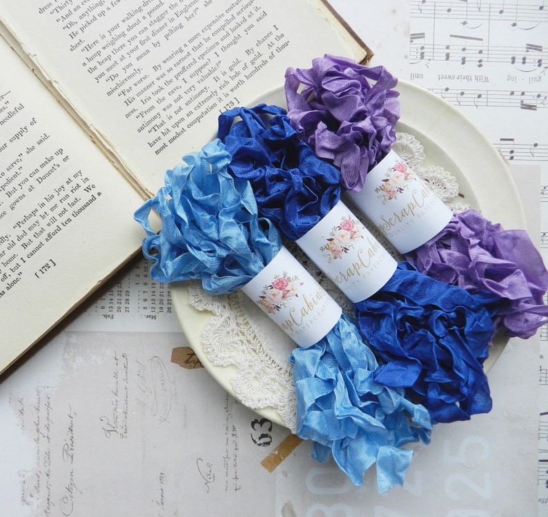 May include: Three spools of ribbon in shades of blue and purple. The ribbon is ruffled and has a satin finish. The spools are labeled "Scrap Cabin".