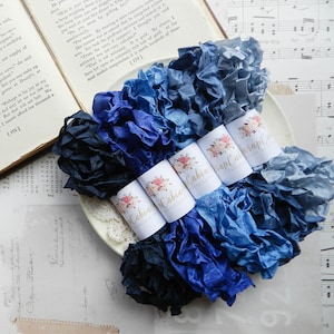 May include: A collection of crinkled silk ribbons in various shades of blue, ranging from deep navy to light slate, bundled together with white paper bands featuring a floral logo and the text Cabin Cabin Scrap Cabin.