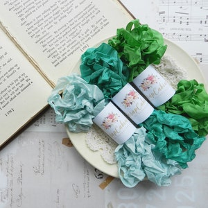 May include: Three spools of ribbon in shades of teal and green, wrapped in white paper with floral designs and the text "Scrap Cabin" printed on each spool.