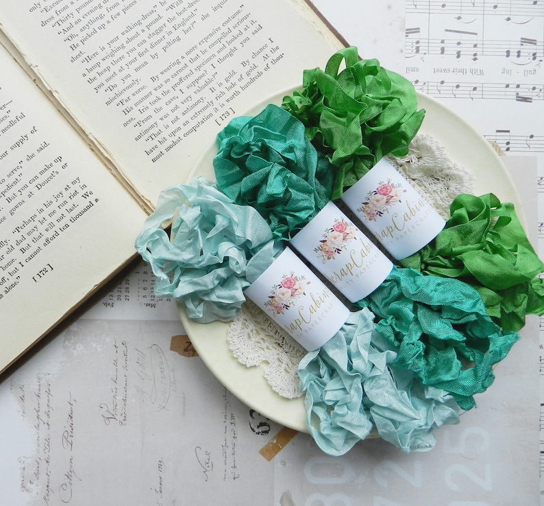 May include: Three spools of ribbon in shades of teal and green, wrapped in white paper with floral designs and the text "Scrap Cabin" printed on each spool.