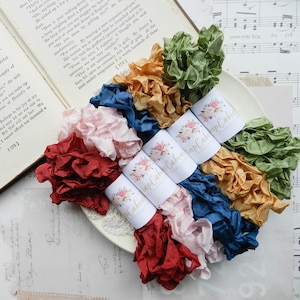 May include: Assortment of colorful, ruffled silk ribbons in shades of red, pink, blue, gold, and green. The ribbons are arranged on a white plate, with floral labels. The background includes pages from a vintage book and sheet music.