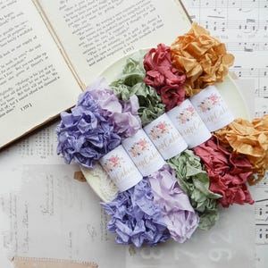 May include: A selection of colourful, ruffled ribbons in shades of purple, pink, green, and gold. Several spools of ribbon are visible, each with a floral design and the text "Cabin Wrap Cabin". The ribbons are arranged on a decorative plate.