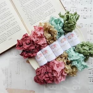 May include: A collection of colorful, ruffled silk ribbons in shades of pink, green, and gold, arranged on a white plate. The ribbons are accompanied by white labels with floral designs and the text "Wrap Cabin". An open book and sheet music are in the background.