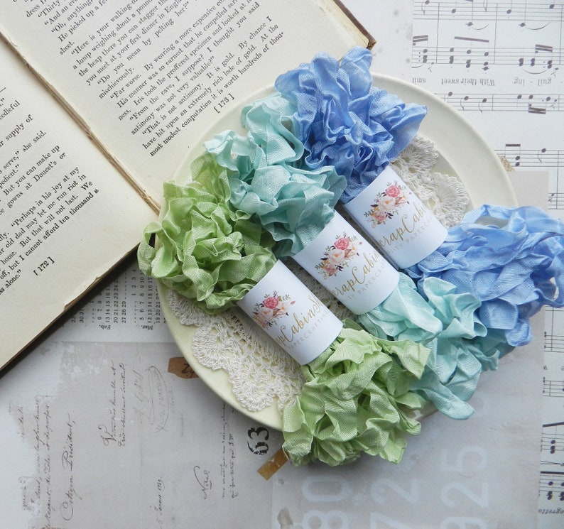 May include: Three spools of ruffled fabric ribbon in shades of light blue, green, and turquoise. Each spool is wrapped with a white label with a floral design and the text "Scrap Cabin Fabrics".