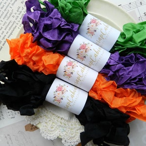 Op de afbeelding: Assortiment van kleurrijke, gerimpelde stofbandrollen met "Happy Cabin Papercraft" labels. De linten zijn zwart, oranje, paars en groen.