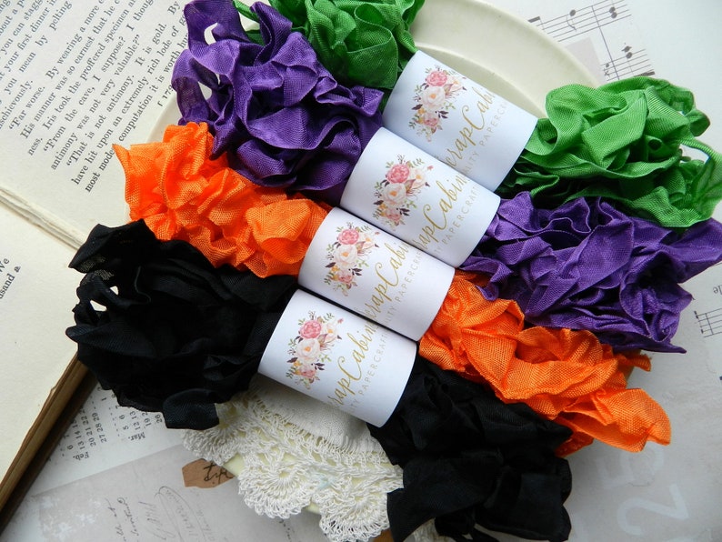 Op de afbeelding: Assortiment van kleurrijke, gerimpelde stofbandrollen met "Happy Cabin Papercraft" labels. De linten zijn zwart, oranje, paars en groen.