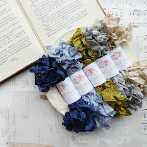 May include: A selection of silk ribbons in various colours, including blue, green, grey, and cream. The ribbons are bundled and labelled with floral designs. An open book and sheet music are in the background.