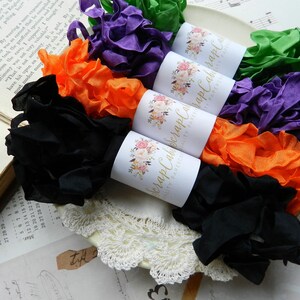 Op de afbeelding: Vijf spoelen met gekleurd lint, elk verpakt in wit papier met een bloemenmotief en de tekst "Scrap Cabin" erop gedrukt. De linten zijn zwart, oranje, paars, groen en een lichtere tint paars.