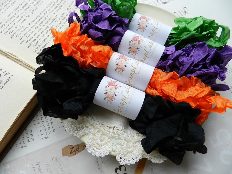 Op de afbeelding: Vijf spoelen met gekleurd lint, elk verpakt in wit papier met een bloemenmotief en de tekst "Scrap Cabin" erop gedrukt. De linten zijn zwart, oranje, paars, groen en een lichtere tint paars.