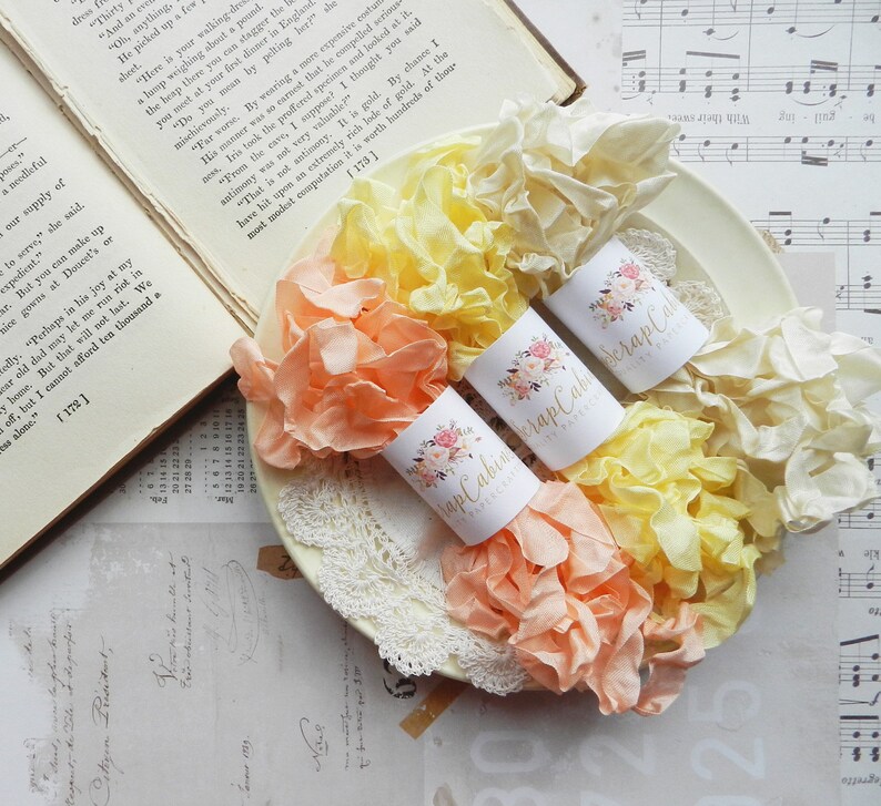May include: Three spools of ruffled ribbon in shades of peach, yellow, and cream. Each spool is wrapped in white paper with a floral design and the text "Scrap Cabin" printed on it.