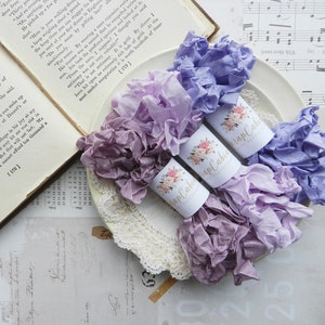 May include: Three spools of purple, lavender, and light blue ribbon with ruffled edges. The spools are wrapped in white paper with a floral design and the text "Scrap Cabin" printed on them.