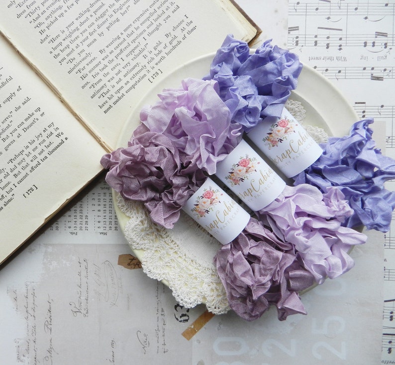 May include: Three spools of purple, lavender, and light blue ribbon with ruffled edges. The spools are wrapped in white paper with a floral design and the text "Scrap Cabin" printed on them.