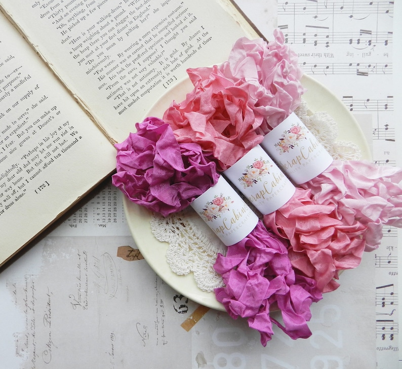 May include: Three spools of pink and purple silk ribbon, each wrapped around a white label with the text "Scrap Cabin" and floral designs.