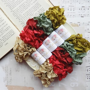 May include: A collection of ruffled fabric ribbon in various colors, including red, gold, green, and olive. The ribbon is bundled and labeled with a floral design and the text "Wrap Cabin Papercraft". The ribbons are arranged on a cream-colored plate.