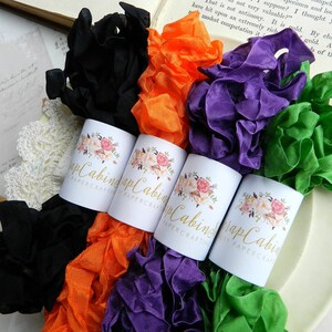 Op de afbeelding: Vier spoelen met gekrulde lint in zwart, oranje, paars en groen. Elke spoel is gelabeld met een bloemmotief en de tekst "Scrap Cabining Papercrafts".