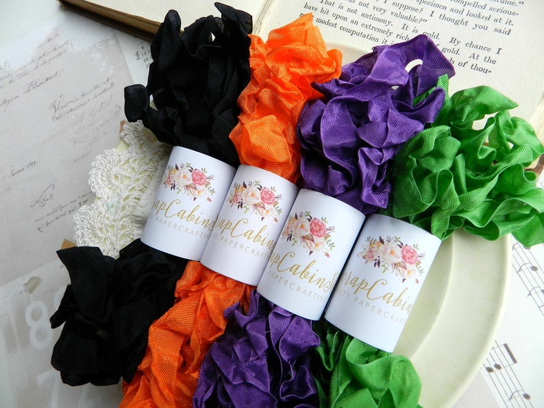 Op de afbeelding: Vier spoelen met gekrulde lint in zwart, oranje, paars en groen. Elke spoel is gelabeld met een bloemmotief en de tekst "Scrap Cabining Papercrafts".