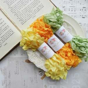 May include: Three spools of ribbon in yellow, orange, and green colors. The ribbon is curled and arranged on a white doily. Each spool has a white label with the text "Wrap Cabin" and a floral design.