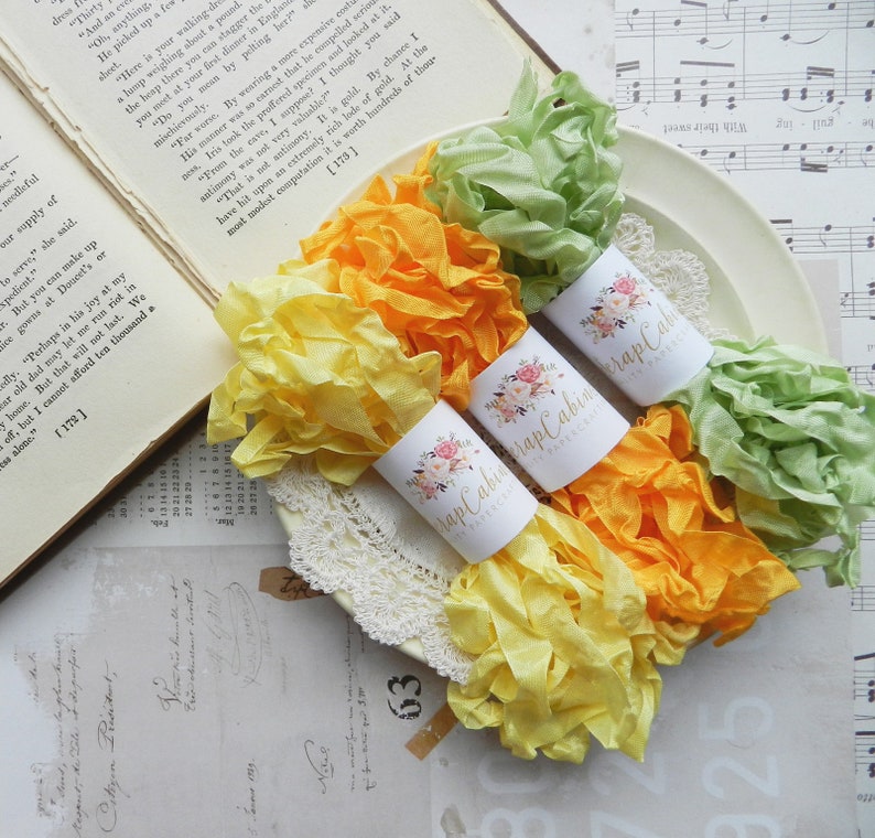 May include: Three spools of ribbon in yellow, orange, and green colors. The ribbon is curled and arranged on a white doily. Each spool has a white label with the text "Wrap Cabin" and a floral design.