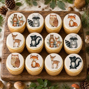 May include: Twelve cupcakes arranged in a grid, each topped with a circular edible topper featuring watercolor illustrations of woodland animals including bears, deer, raccoons, owls, foxes, and skunks, set on a wooden board.