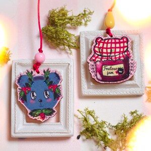 May include: Two decorative ornaments. One features a blue bear with a berry wreath, the other a jar of jam with the text "Fruity Jam". Both are set in white frames and suspended by red cords with pink beads.