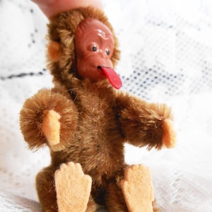 May include: A brown plush monkey toy with a red tongue sticking out. The monkey has a brown fur coat and a light brown face. The monkey is sitting with its legs crossed.