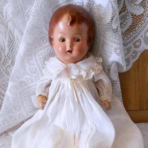 May include: A vintage composition doll with reddish-brown hair and painted facial features, wearing a long, white, sheer gown featuring delicate lace trim, a pleated front panel, a Peter Pan collar, and a thin white ribbon tied at the neckline.